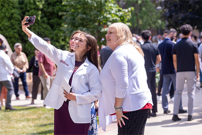 2025_CMEDWhiteCoat_700x467 Three women are taking a selfie outdoors during a sunny day. People and greenery are visible in the background.