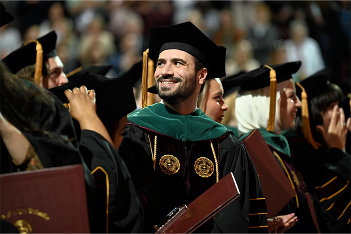 2025_CMEDCommencement_700x467 A group of graduates in black gowns and caps stand during a ceremony. One person in the center, smiling broadly, wears a green academic hood and has two medallions on their gown. Others around them also wear gowns and hold diploma folders.
