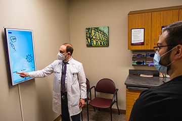 A male doctor in a face mask points to a display board to show his male patient some vaccine information.