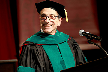 Man in green and black doctoral graduation regalia stands at a podium smiling.