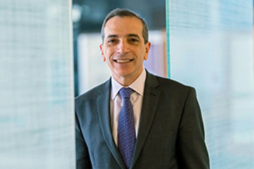 Man in a dark suit with a white shirt and blue tie leans against a glass wall, smiling at the camera.