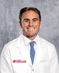 A man with short, dark hair wearing a white collared shirt and blue tie wears a medical white coat and smiles for a headshot.