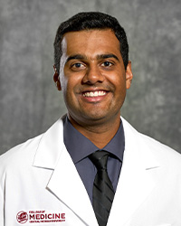 A man with short, dark hair wearing a dark gray collared shirt and black tie wears a medical white coat and smiles for a headshot.
