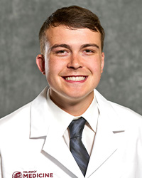 A man with short, dark hair wearing a white collared shirt and blue/gray tie wears a medical white coat and smiles for a headshot.