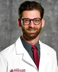A man with short, dark hair, mustache and beard wearing a dark gray collared shirt and maroon tie wears a medical white coat and smiles for a headshot.