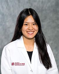 A woman with long, black hair and a black blouse wears a medical white coat and smiles for a headshot.