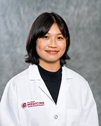 Nguyen, Ellie 200 x 250 web A woman with medium-length brown hair wearing a black shirt and a medical white coat as she smiles at the camera.