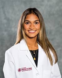 Sanku, Deepti 200 x 250 web A woman with long blond brown hair wearing a blue shirt and a medical white coat as she smiles at the camera.