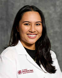 A woman with long, dark hair and a black blouse wears a medical white coat and smiles for a headshot.