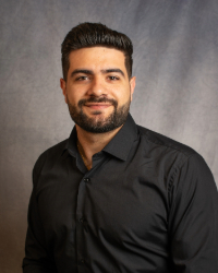 A man with short dark hair with mustache and beard wearing a dark collared shirt.