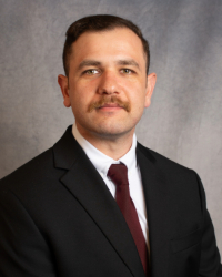 A man with dark and mustache wearing a dark suit with a white collared shirt and maroon tie.