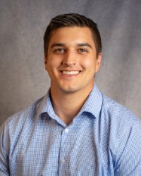 A man in a blue shirt smiles for a professional headshot in front of a grey background.