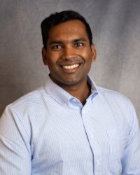 A man in a blue button up shirt smiles for a professional headshot in front of a grey background.