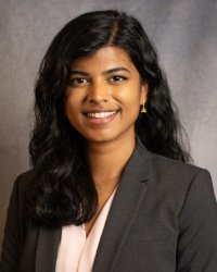 A woman in a dark suit coat and light blouse smiling for a headshot in front of a grey background.