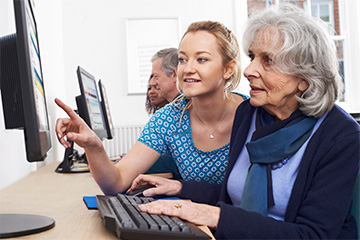 A young woman helps a female senior citizen learn how to use the internet on a desktop computer.