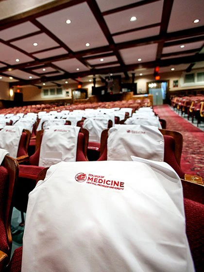 White coats with the CMU College of Medicine logo rest on the backs of chairs in a large auditorium.