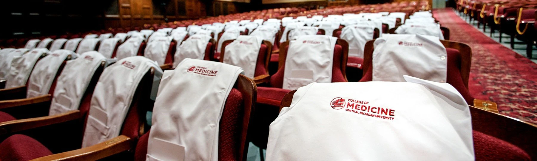 White coats with the CMU College of Medicine logo rest on the backs of chairs in a large auditorium.