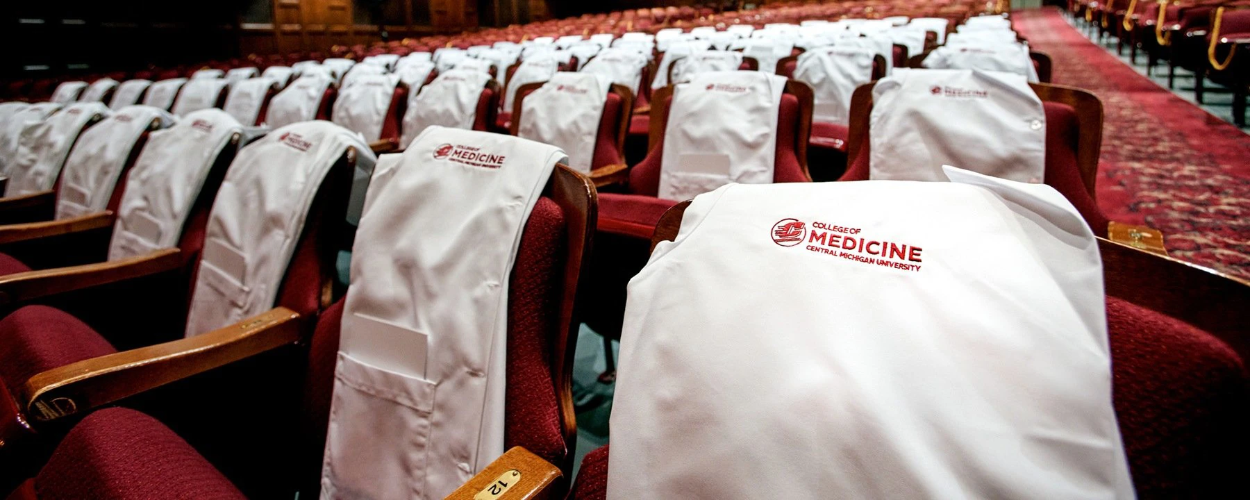 White coats with the CMU College of Medicine logo rest on the backs of chairs in a large auditorium.