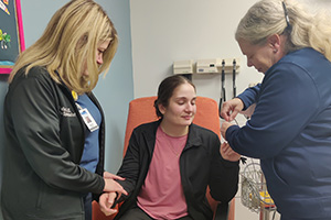 A woman seated in a clinical exam room is assisted by two, female health care providers.