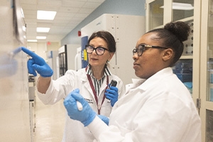 Two women wearing lab coats and gloves discussing data on a whiteboard in a science lab.