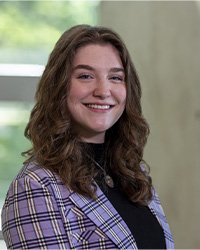 A woman with long brown hair wearing a black shirt and plaid jacket smiles at the camera.