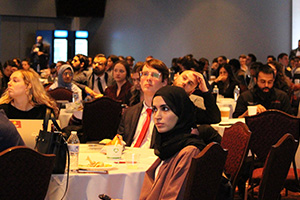 A diverse audience of professionals and students sits at round tables during a research symposium, focused on a presentation.