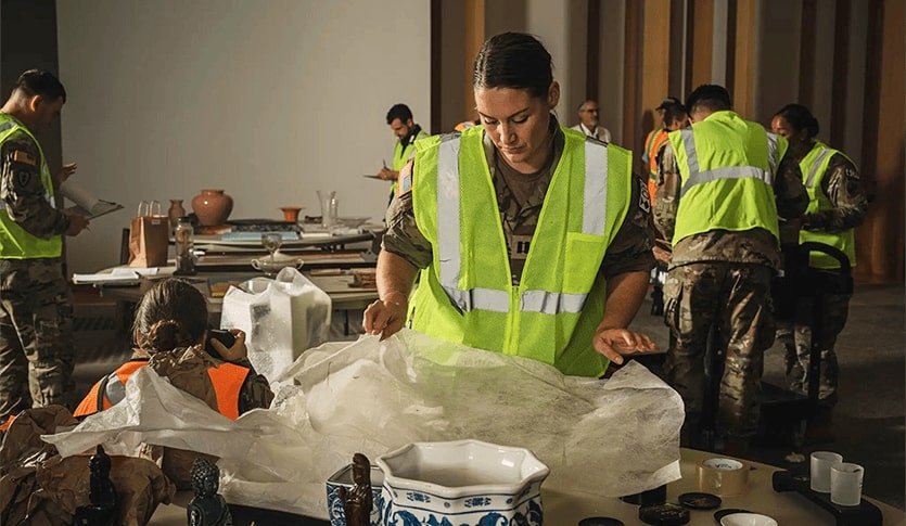 Jessica Wagner storing artifacts for a museum while serving in the military. She is wearing her fatigues and a yellow vest. There are several ceramic artifacts on a table in front of her.