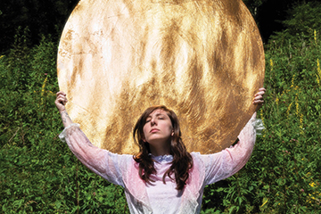 20260202_Events_Owensby_003_360x240 Artist Danielle Owensby is holding a large golden disc up behind and above her head. The sun is reflecting off of it. There is a wall of greeenery behind her.