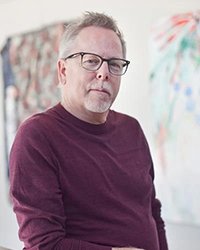 Headshot of Clyde Sheets standing in front of multicolored paintings hanging on the wall behind him.