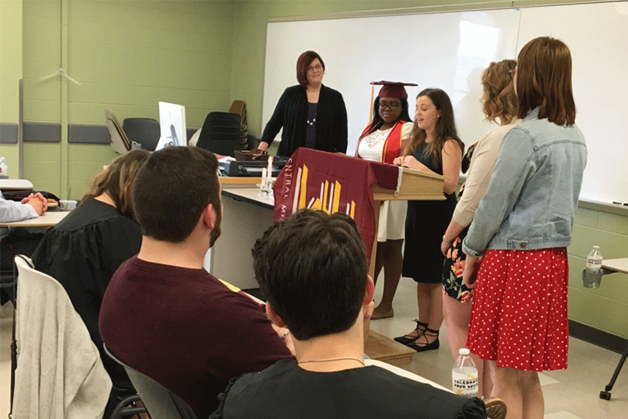 Students standing in a classroom, while some are sitting at their desk as they listen to those standing and speaking at a podium.