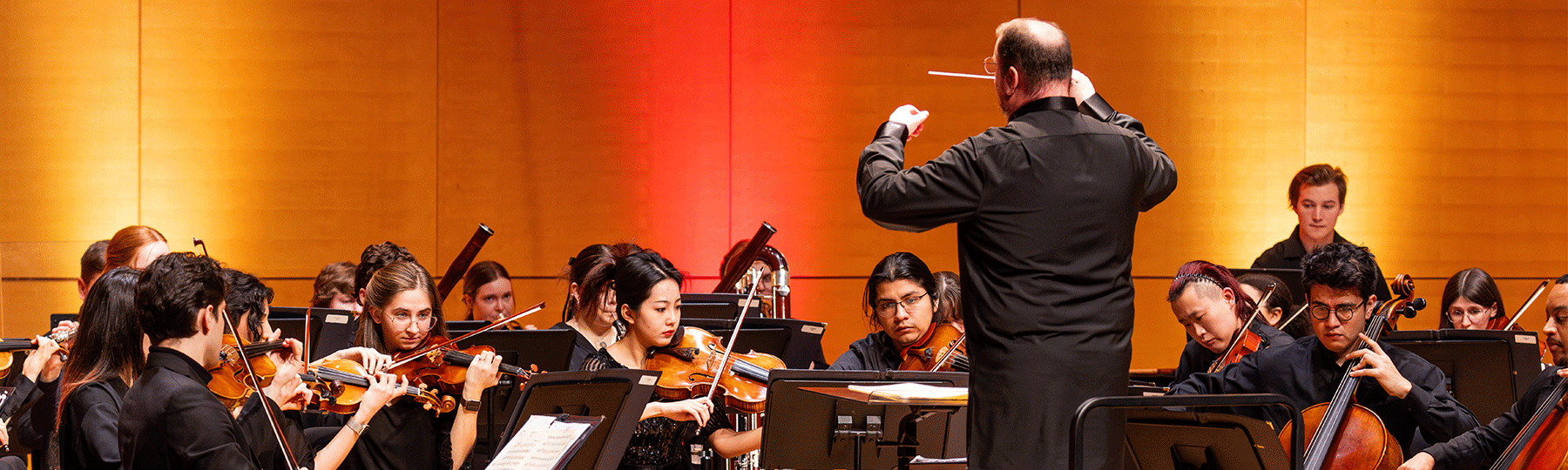 A conductor stands on stage while a group of students play instruments on stage during a performance.