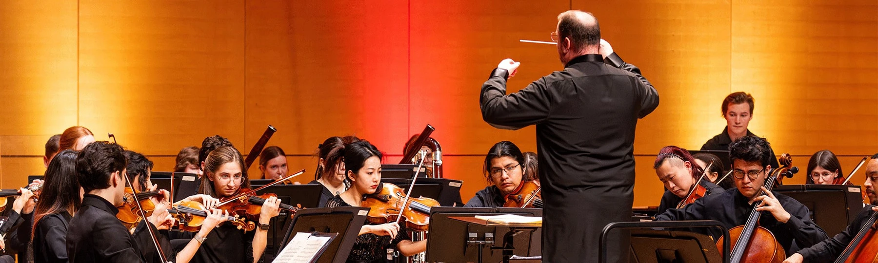 A conductor stands on stage while a group of students play instruments on stage during a performance.