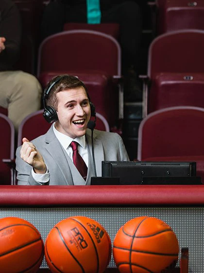 A male student wearing a headset broadcasts a basketball game.