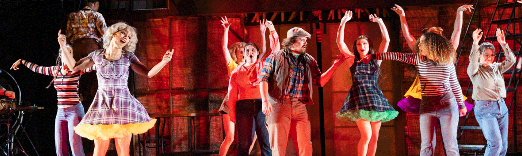 A group of theatre students on stage as they are acting during a performance, there is red stage lighting behind them, and some performers are wearing plaid dresses.