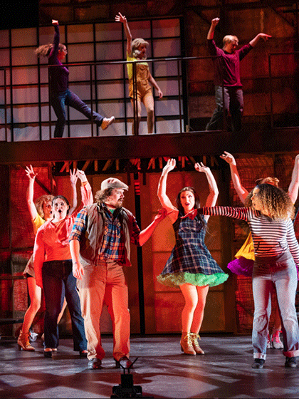 A group of theatre students on stage as they are acting during a performance, there is red stage lighting behind them, and some performers are wearing plaid dresses.