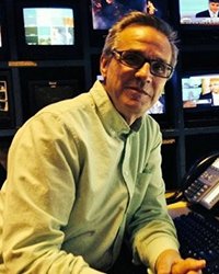 Headshot of Tim Roberts. He is in the tv control room. You can see several screens behind him.