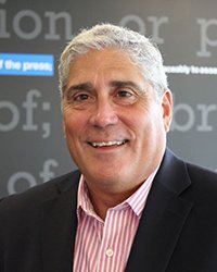 Headshot of Ed Fernandez set in front of newsroom media screens.