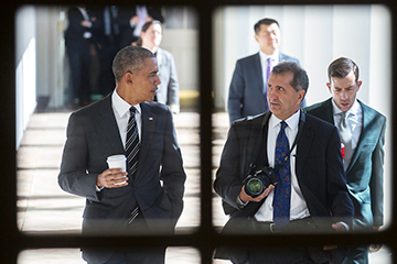 Image shot through a window frame of Pete Souza with President Obama. Pete is holding a camera. The President is holding a cup of coffee. They are walking down a corridor.