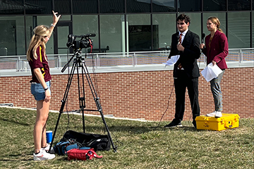 Sarah Kelly working as cameraperson at a home football game. Two fellow students are working as sports journalists in front of her camera.