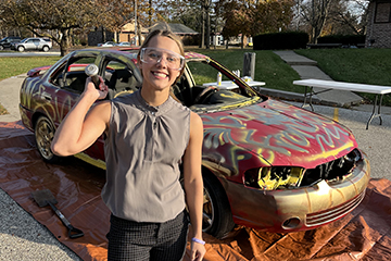 Ella Pierzecki wearing safety glasses with sledge hammer in hand, standing in front of the car that will be smashed as a homecoming tradition.