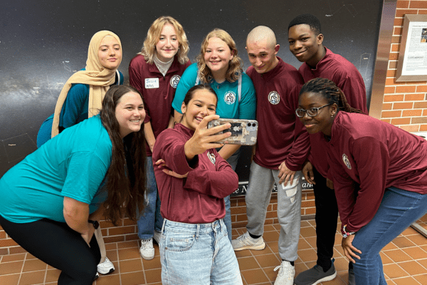 A group of seven middle schoolers cluster around a cell phone with their teacher for a selfie. They are smiling.