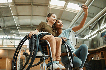 Girl in a wheelchair with another girl taking a selfie.