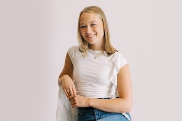 A woman with shoulder-length blonde hair and wearing a white shirt is sitting on a chair in front of a white background.