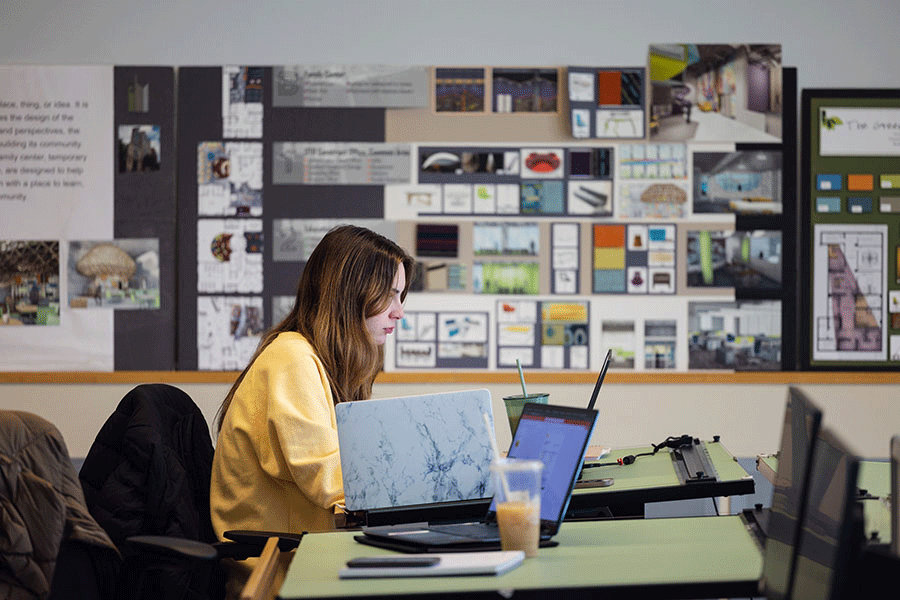 A student sitting at a desk using a laptop computer to complete interior design class assignments.