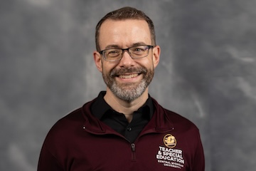 Professional headshot of Troy Hicks against a grey background.