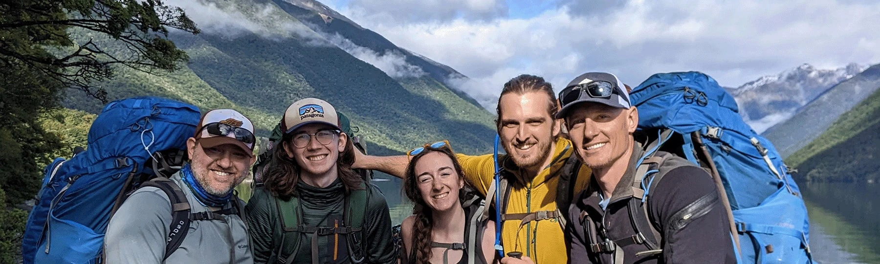 Five people standing outside, in front of a large green mountain with the blue sky in the background. Everyone is wearing hiking gear such as hats, glasses, and backpacks.