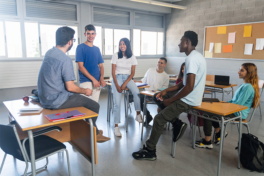 A group of students sit in chairs in a circle as a school counselor speaks to them. They are in a classroom setting.