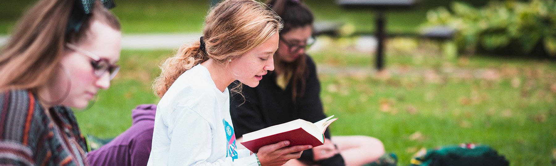Student seated on the grass holds a book in her hands and reads while other students sit nearby to listen.