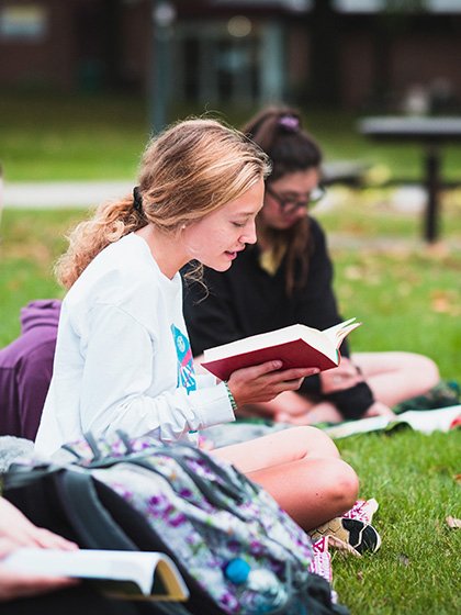 Student seated on the grass holds a book in her hands and reads while other students sit nearby to listen.