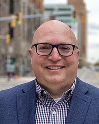 Closeup image of John Sellek wearing glasses and a blue suit jacket standing near a street.